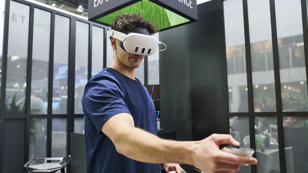 A man with VR glasses and an outstretched controller stands in a trade fair stand, a black cube with the inscription 