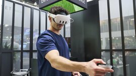 A man with VR glasses and an outstretched controller stands in a trade fair stand, a black cube with the inscription 