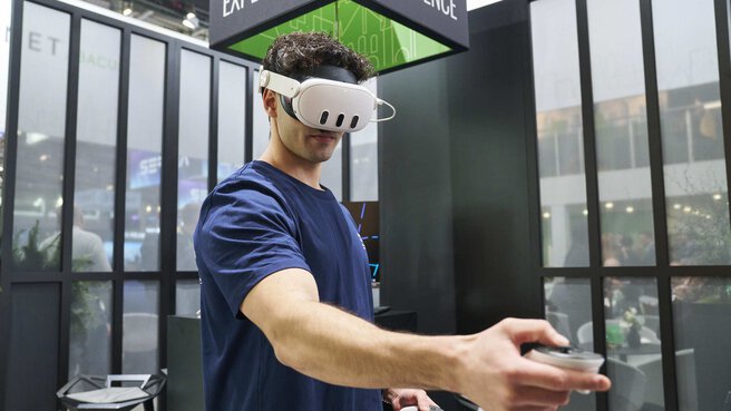A man with VR glasses and an outstretched controller stands in a trade fair stand, a black cube with the inscription 