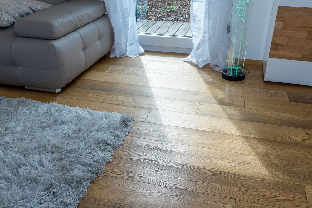 Ein Sonnenstrahl scheint auf einen Holzboden neben einem grauen Sofa und einem zotteligen Teppich, in der Nähe eines Vorhangs und einer Glastür.