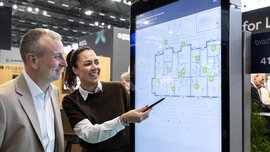Zwei Personen betrachten und besprechen einen digitalen Grundriss, der auf einem großen interaktiven Bildschirm bei einer Veranstaltung angezeigt wird.
