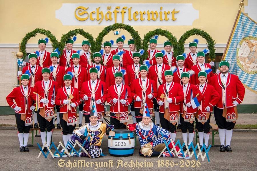 Gruppenfoto  der Schäfflerzunft Aschheim von 1886-2026. Die Personen tragen rote Uniformjacken, schwarze Hosen und grüne Hüte. Zwei sind als Harlekine verkleidet.