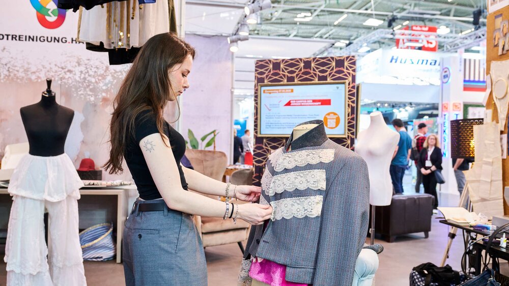 A young woman presenting sustainable fashion at a trade fair stand, adjusting a grey blazer with lace trim on a mannequin.
