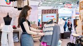 Eine junge Frau präsentiert an einem Messestand nachhaltige Mode und passt ein graues Sakko mit Spitzenbesatz an einer Schneiderpuppe an.