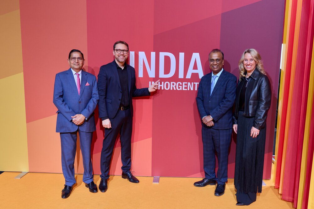 Vier Personen stehen lächelnd und in Abendgarderobe gekleidet vor einer rot-orangefarbenen Wand mit der Aufschrift „INDIA INHORGENTA“.
