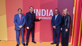 Vier Personen stehen lächelnd und in Abendgarderobe gekleidet vor einer rot-orangefarbenen Wand mit der Aufschrift „INDIA INHORGENTA“.