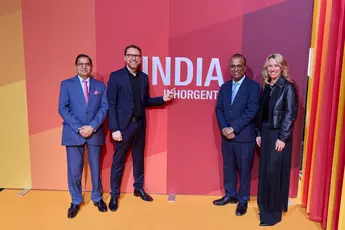 Four people stand smiling and dressed in evening wear in front of a red-orange coloured wall with the inscription ‘INDIA INHORGENTA’.