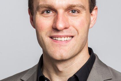 Smiling person in a light gray blazer and a black shirt against a plain background.