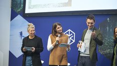 Three smiling people with microphones on a trade fair stage.