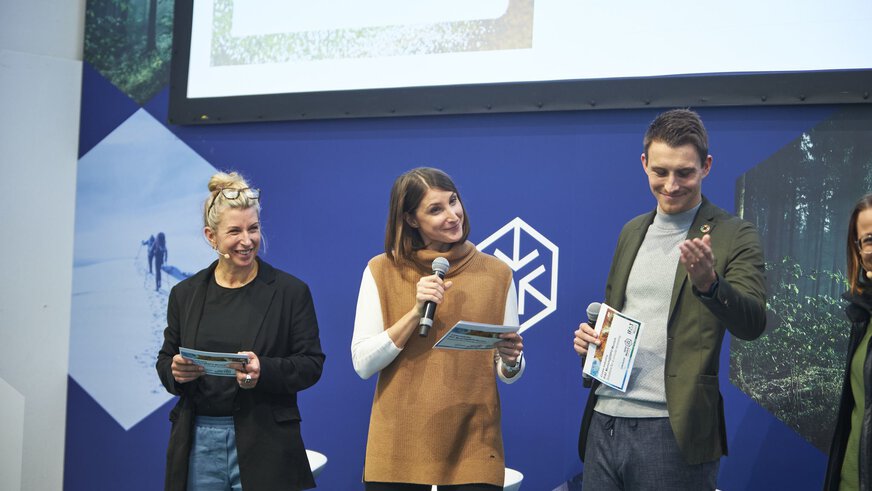 Three smiling people with microphones on a trade fair stage.