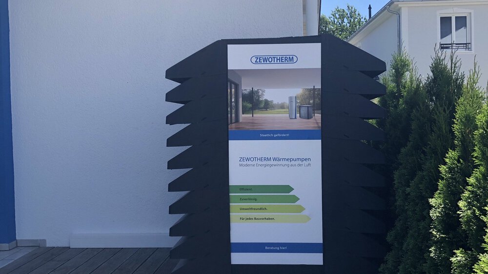 Außendisplay mit einer Werbung für ZEWOTHERM Wärmepumpen vor einem Musterhaus.