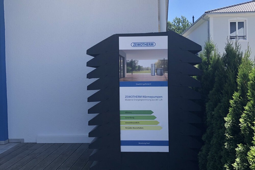Außendisplay mit einer Werbung für ZEWOTHERM Wärmepumpen vor einem Musterhaus.