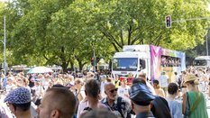 Eine große Menschenmenge feiert im Freien neben einem mit Regenbogenfarben geschmückten Lastwagen unter grünen Bäumen.