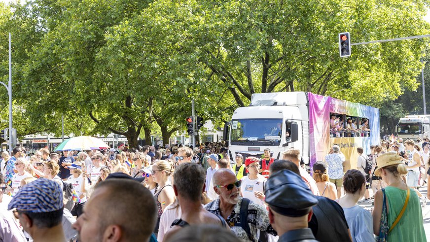 Eine große Menschenmenge feiert im Freien neben einem mit Regenbogenfarben geschmückten Lastwagen unter grünen Bäumen.