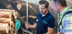 An exhibitor and an INTERFORST visitor look at a tablet together. Stacks of wood lie next to them.