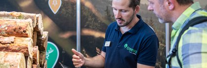 Ein Aussteller und ein Besucher INTERFORST blicken gemeinsam auf ein Tablet. Neben ihnen liegt gestapeltes Holz.