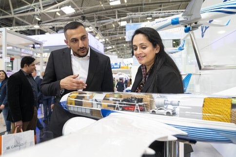 Two people look smilingly at the model of an airplane with a transparent cargo hold in which various goods are displayed.