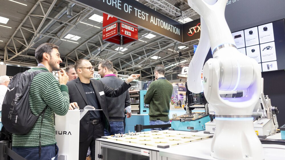 Mehrere Besucher stehen an einem Stand auf der productronica und betrachten die Ausstellungsstücke, unter anderem einen weißen Roboterarm.