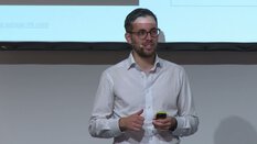 Ein Mann mit Brille und weißem Hemd spricht ins Mikrofon und hält einen Clicker in der Hand.