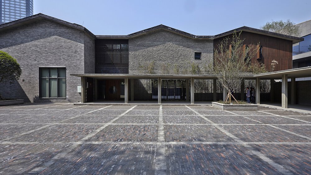 Modernes Gebäude mit grauer Backsteinfassade, schrägen Dächern und einem großen offenen Innenhof unter klarem Himmel.
