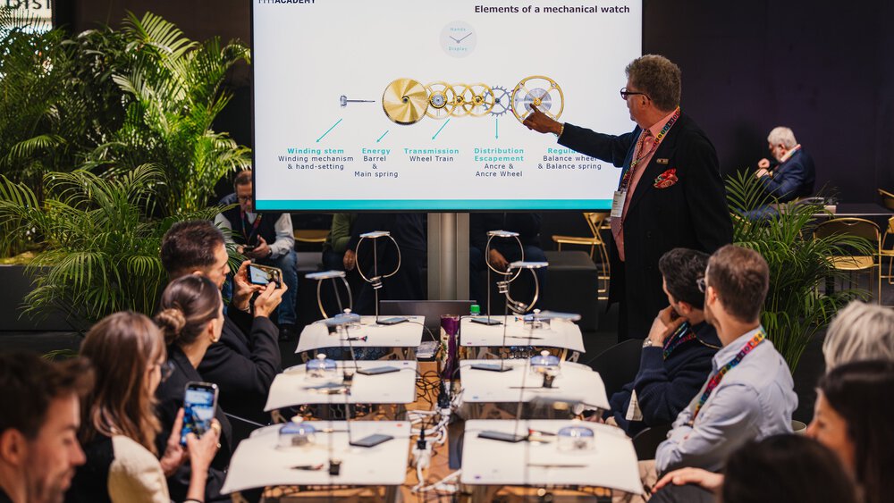 A man stands in front of a screen and explains the individual elements of a mechanical watch to an interested audience.