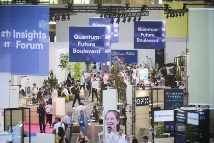 Blick auf eine Messehalle mit “Quantum Future Boulevard“-Bannern, zahlreichen Ständen und Besuchern.