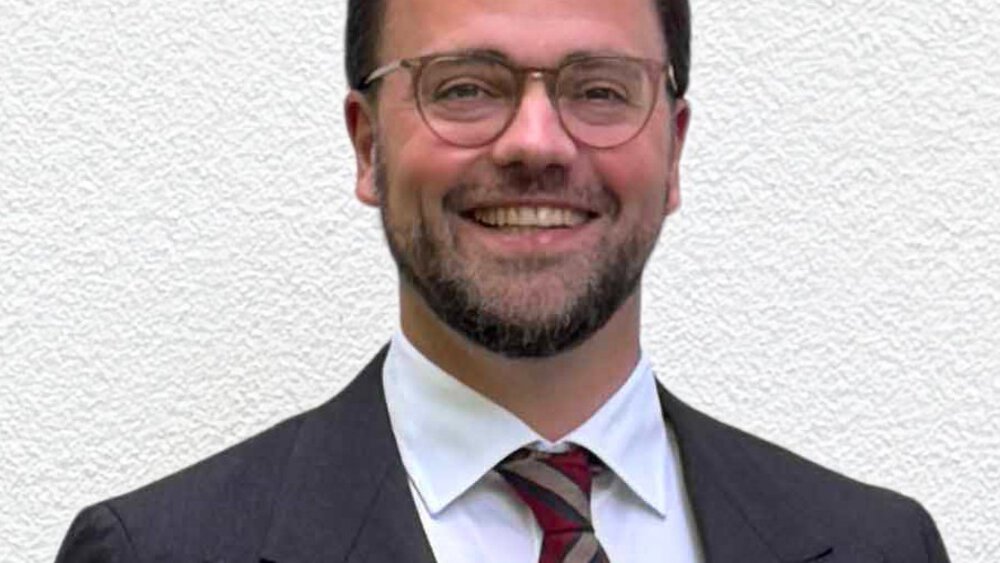 A man with short brown hair, glasses, a beard, and wearing a black suit with a red striped tie smiles.