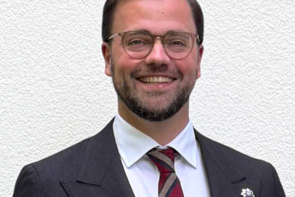 Ein Mann mit kurzen braunen Haaren, einer Brille, Bart und in einem schwarzen Anzug mit einer rot gestreiften Krawatte lächelt.