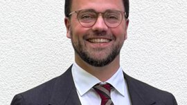 Ein Mann mit kurzen braunen Haaren, einer Brille, Bart und in einem schwarzen Anzug mit einer rot gestreiften Krawatte lächelt.