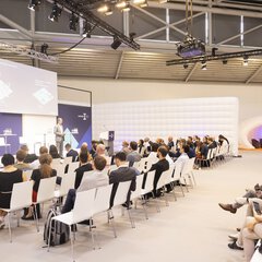 Stage of the munich_i with a full audience on white chairs and a speaker in a gray suit.