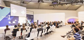 Bühne der munich_i mit vollem Publikum auf weißen Stühlen und einem Sprecher in einem grauen Anzug.