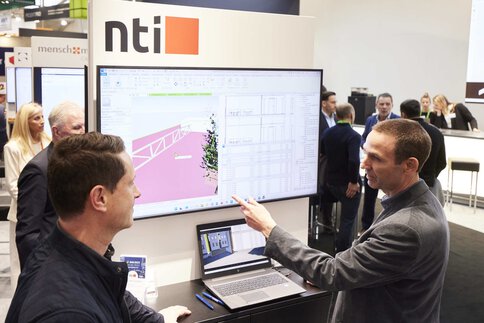 Zwei Männer diskutieren an einem Messestand vor einem Bildschirm, auf dem ein Designplan angezeigt wird. Im Hintergrund stehen weitere Personen.