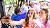 Zwei Frauen, die bei einer farbenfrohen Veranstaltung im Freien für ein Selfie lächeln, umgeben von Menschen in festlicher Kleidung.