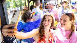 Two women smiling for a selfie at a colorful outdoor event, surrounded by people in festive attire.