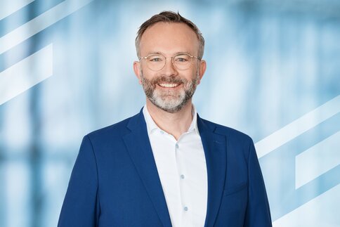 Lächelnder Mann mit Brille und Bart in einer blauen Anzugsjacke, stehend vor einem blauen geometrischen Hintergrund.