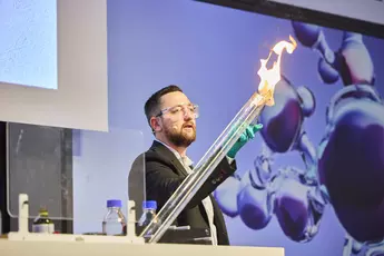 A man in a suit with protective goggles and laboratory gloves stands on a stage and carries out an experiment. He is holding something burning in front of a long transparent tube. In the background you can see an analytica Keyviusal.