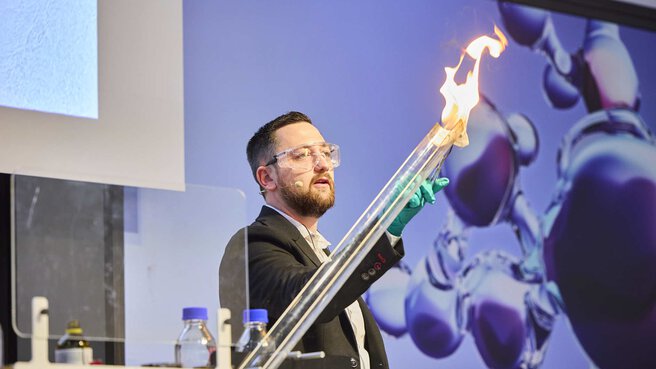 A man in a suit with protective goggles and laboratory gloves stands on a stage and carries out an experiment. He is holding something burning in front of a long transparent tube. In the background you can see an analytica Keyviusal.