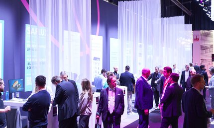  Zahlreiche Menschen stehen an einer Reihe Messestände, die durch helle, halbtransparente Vorhänge abgetrennt sind. Die Umgebung ist blau und violett beleuchtet.