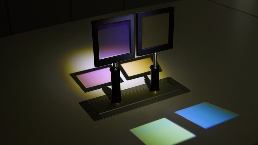 Eine optische Experimentieranordnung mit zwei rechteckigen Farbfiltern in schwarzen Rahmen, die auf einer Tischhalterung montiert sind. Durch die Filter fällt Licht, das auf der Tischoberfläche verschiedenfarbige Projektionen erzeugt, darunter Gelb, Blau, Grün und Violett.
