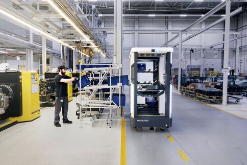 A black and white robot cabinet on castors stands next to a worker in a large warehouse with numerous machines, shelves and white support beams.