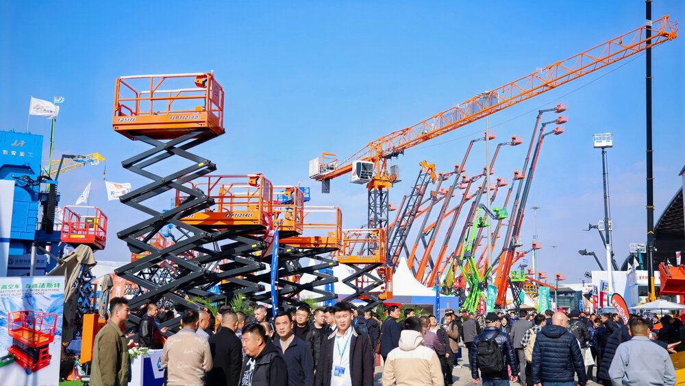 Crowds of people are walking around among large construction machines, cranes, and scissor lifts at an outdoor trade show.