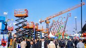 Crowds of people are walking around among large construction machines, cranes, and scissor lifts at an outdoor trade show.