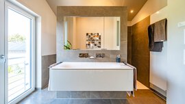 Modernes Badezimmer mit großem Spiegel, hängendem Waschbecken, Handtüchern, begehbarer Dusche und einem raumhohen Fenster, das für Tageslicht sorgt.