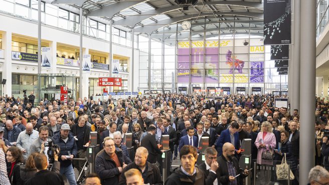Eine große Menschenmenge geht durch einen belebten Eingangsbereich der Messe München, mit Bannern und Werbeanzeigen über den Köpfen und an den Wänden.