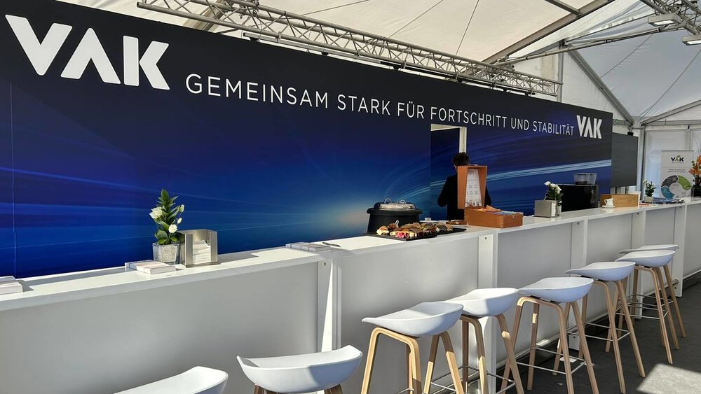 Messestand von VAK auf der IFAT Munich mit moderner Bar-Theke, Sitzhockern und Branding ‚Gemeinsam stark für Fortschritt und Stabilität‘ unter einem hellen Zeltdach.