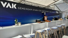 Messestand von VAK auf der IFAT Munich mit moderner Bar-Theke, Sitzhockern und Branding ‚Gemeinsam stark für Fortschritt und Stabilität‘ unter einem hellen Zeltdach.
