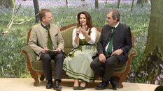 Two men, with a woman between them, sit in traditional clothing with microphones on a green couch at INTERFORST.