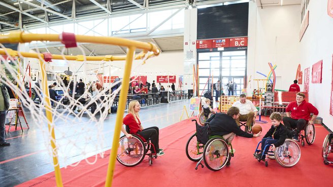 Menschen im Rollstuhl spielen Basketball auf einem roten Spielfeld in einer Messehalle mit verschiedenen Sport- und Spielstationen.