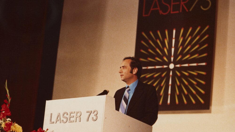Man in suit and striped tie speaks at a podium. A sign with the 