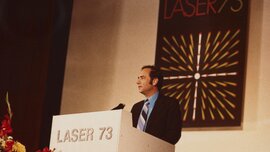 Man in suit and striped tie speaks at a podium. A sign with the 